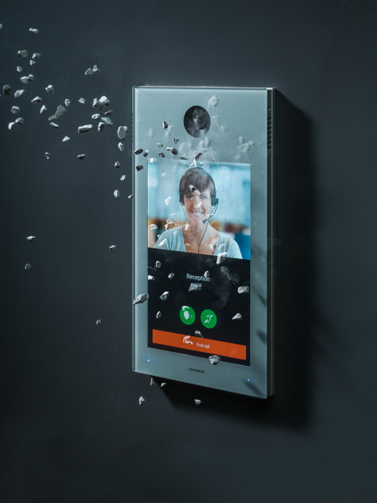 Robustes Video-Türsprechsystem mit Schutzglas bei Beschädigung Ein Wandgerät mit Kamera und Touchscreen zeigt eine Videounterhaltung mit einer Frau. Das Gerät wird von aufprallenden Objekten getroffen, ohne sichtbar zu zerbrechen.