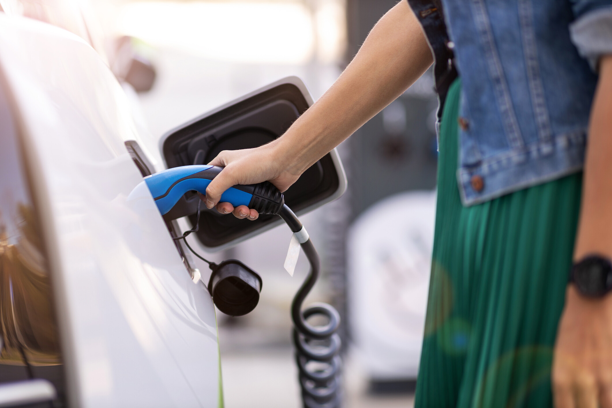 Ladevorgang eines Elektroautos Eine Person steckt ein blaues Ladekabel in die Ladebuchse eines weißen Elektroautos. Der Ladevorgang findet an einer modernen Ladestation statt.