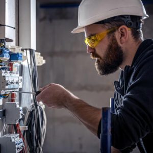Elektro-Installateur mit Schutzbrille und weißem Helm arbeitet am Elektroverteiler